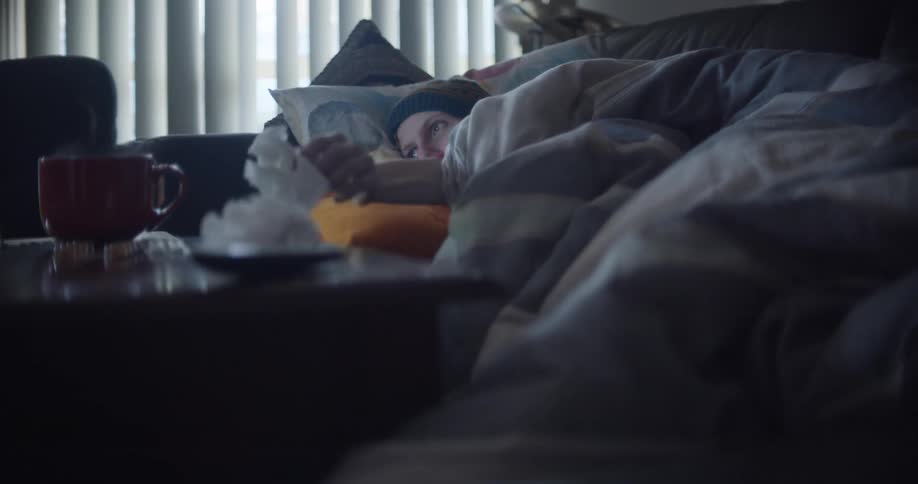 An ill man lies on the couch at home and blows his nose as he watches his computer screen
