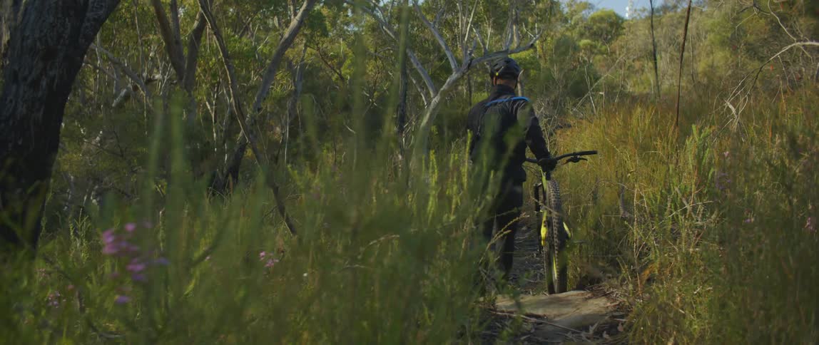 A man wheels his mountain bike along a narrow trail through tall grass toward trees