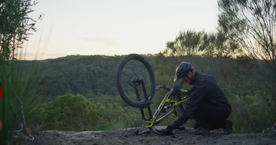Man crouches near upended bicycle examining wheel with forest covered hill sitting under red tinted horizon in background
