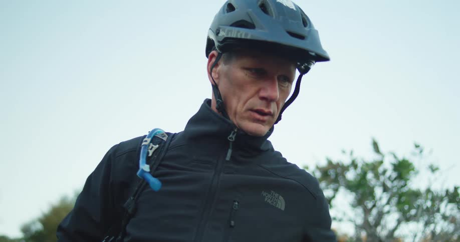 A man outside with a helmet on lifts a backpack on to his back