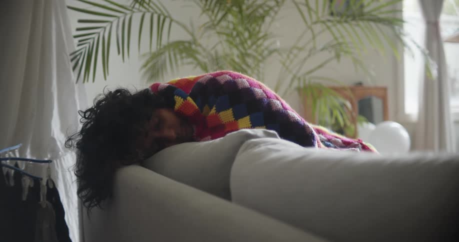 A woman covered in a blanket lies on her couch and sleeps at home