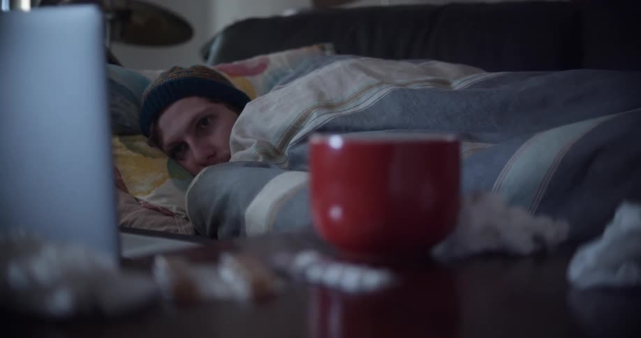A man rests under a blanket on a sofa while watching the display on his laptop