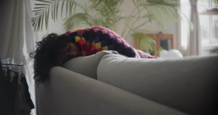 A woman rests on a sofa with a colorful blanket wrapped around her during the day