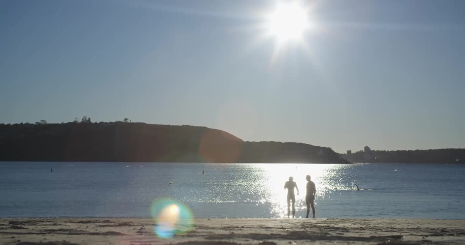 2 people stand at the edge of the water talking in the sunshine