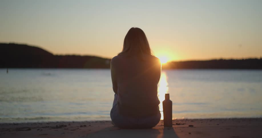 A woman sits on a lakeside beach with a drink container beside her