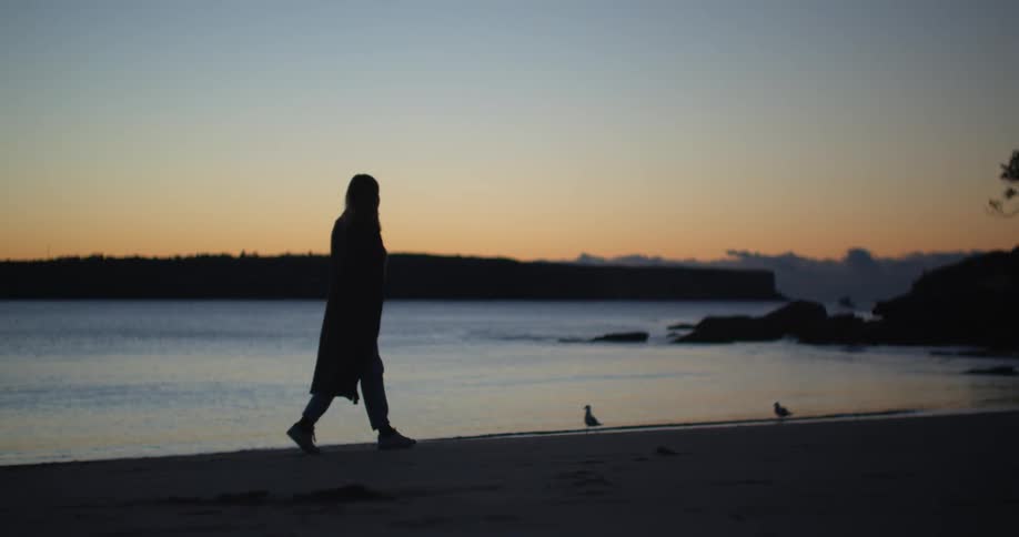 A woman leisurely walks across a beach while gazing out across the ocean at sunset