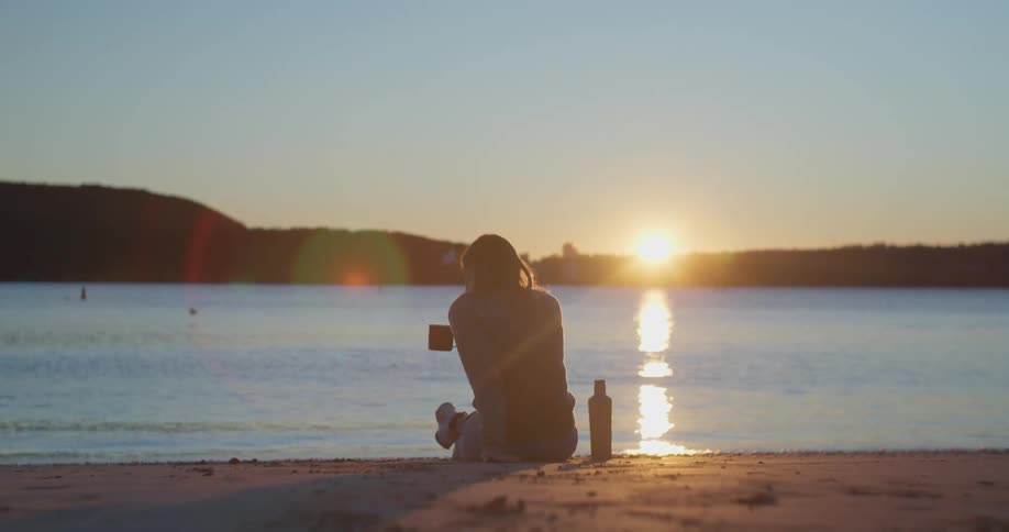A person sitting by a body of water with the sunset the sky and clouds in the background