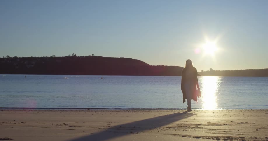 A woman is walking on a beach with the sun shining on her back