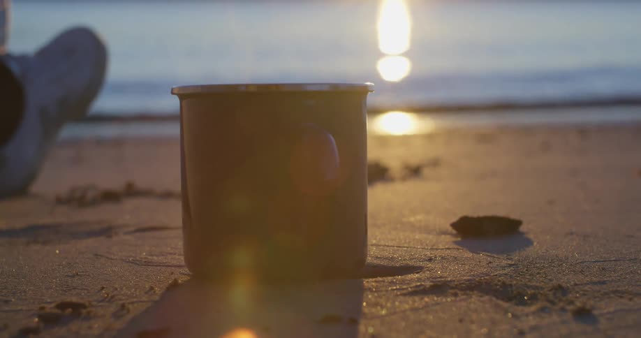 A person sits on the beach with a metal coffee cup and watches the sunset