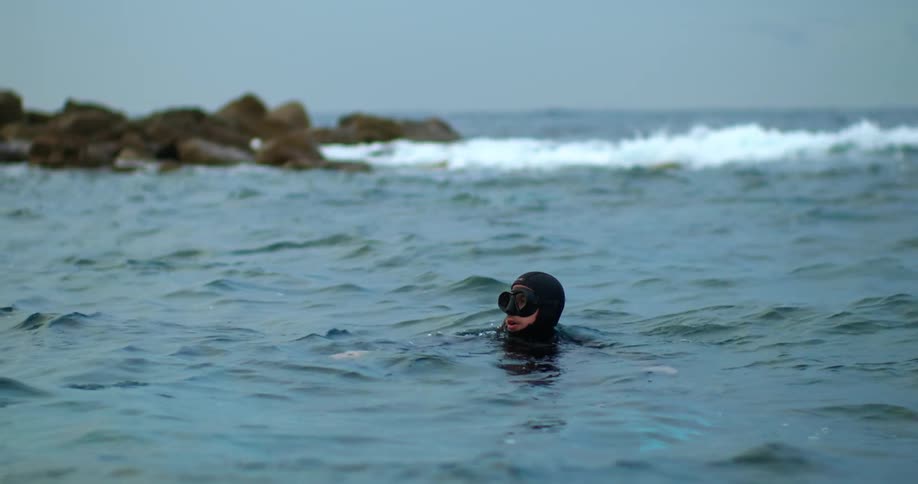 Snorkeling man sinks below water as waves crash against rocks in the background
