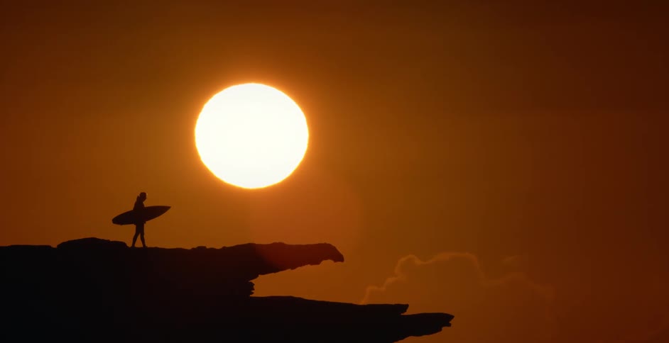 A silhouette of a person holding a surfboard with the sun the sky and clouds in the background