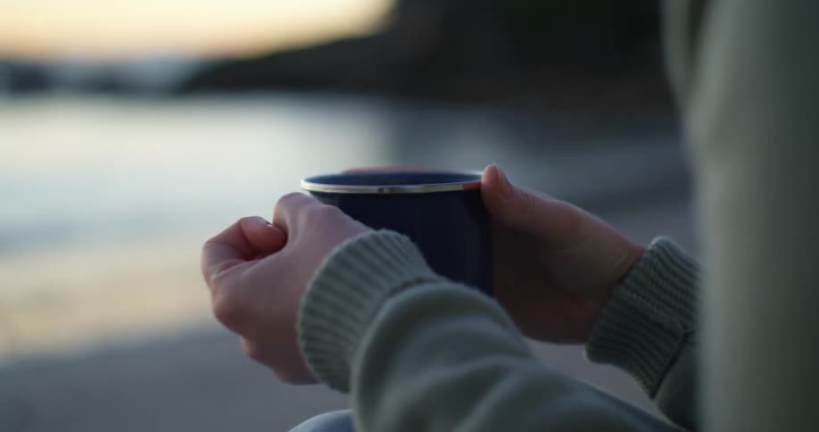 A woman holds a mug while sitting on the beach at sunset