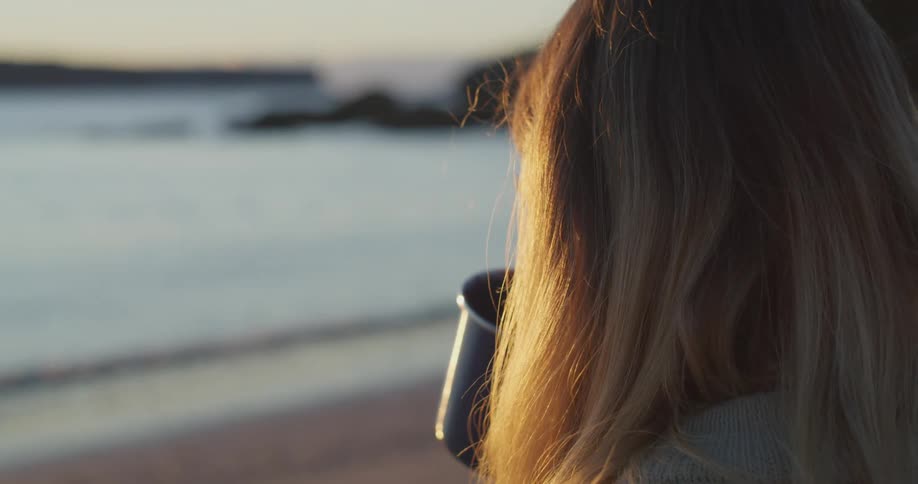 A woman sips coffee from a mug as she sits on the beach