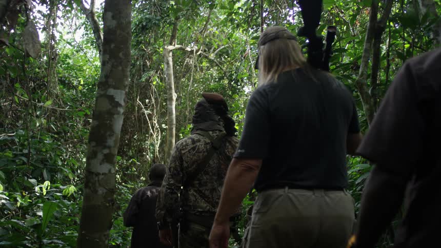 Several men walking slowly through a forest area carrying their guns