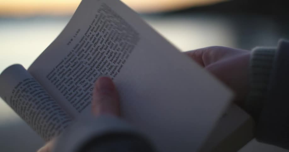 A person with a sweater reading a book with the sky and clouds in the background
