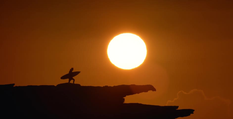 Silhouette of a person walking on a rocky ledge holding a surfboard with a bright sun in a red sky