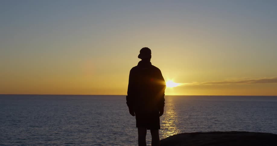 Sun sitting just above horizon shines across ocean to silhouette man standing on shore