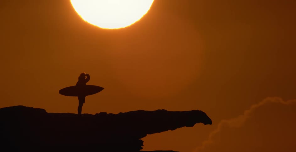Silhouette of a person standing on a rocky ledge holding a surfboard with a bright sun in a red sky