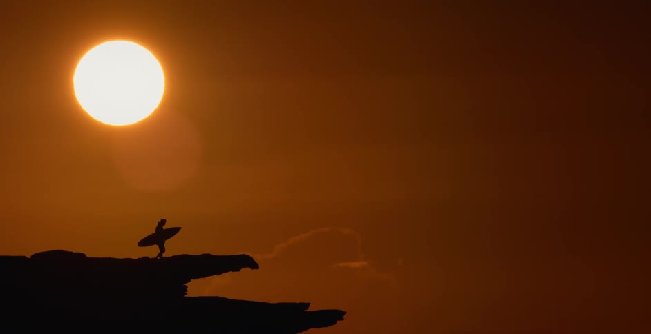 A person is carrying a surfboard across rocks with a red sky and bright sun in the distance