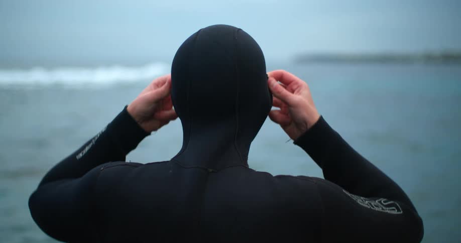 A man standing on the shore pulls the hood of his wet suit on to his head