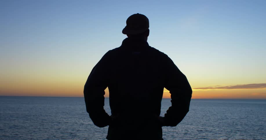 A silhouette of a man standing by the coast with the ocean sky and clouds in the background