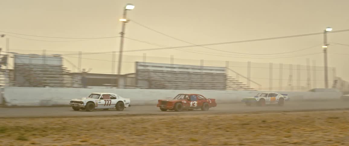 Race cars race around a dirt racing track on a cloudy evening