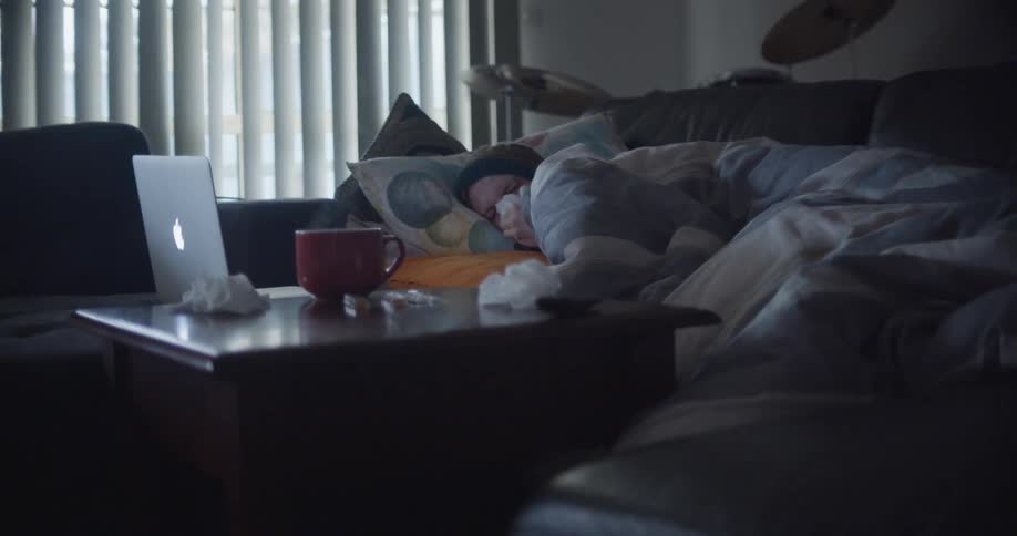 An ill man huddles under a blanket and blows his nose while lying down on his couch and using his laptop