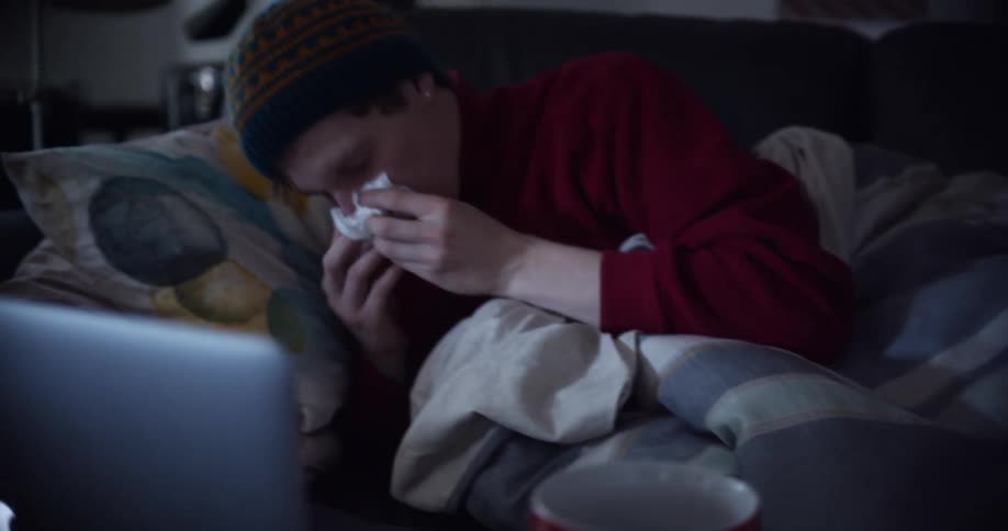 A sick man blows his nose while laying on the couch