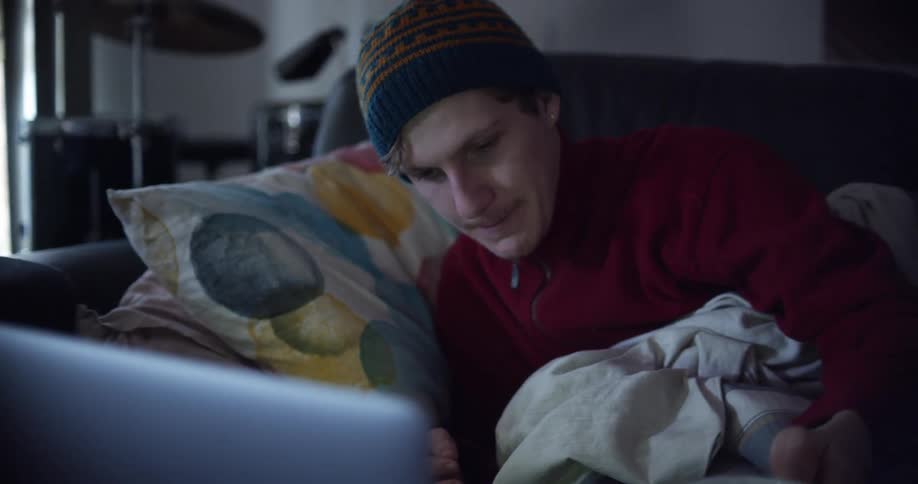 Man wearing a toque drinks a beverage from a large coffee cup then lays on a sofa with pillows and a blanket
