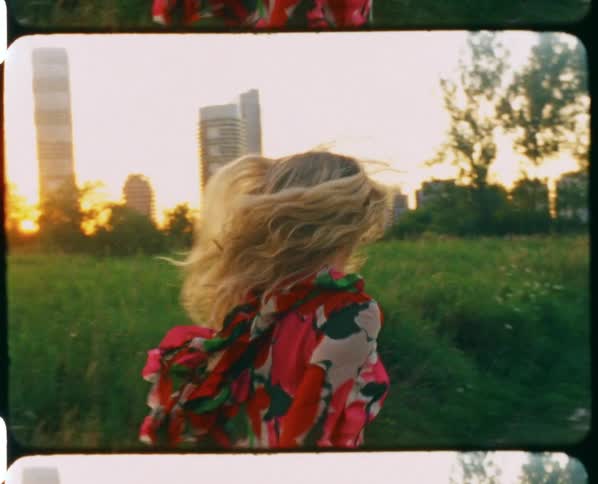 A woman with a dress running in a grassy field with trees buildings the sky and clouds in the background
