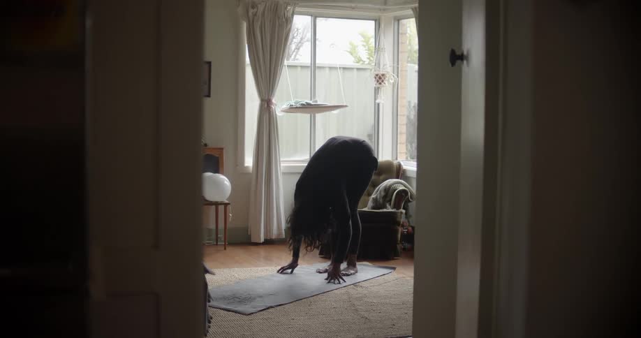 A woman stretches her arms up and bends down while doing yoga in her living room