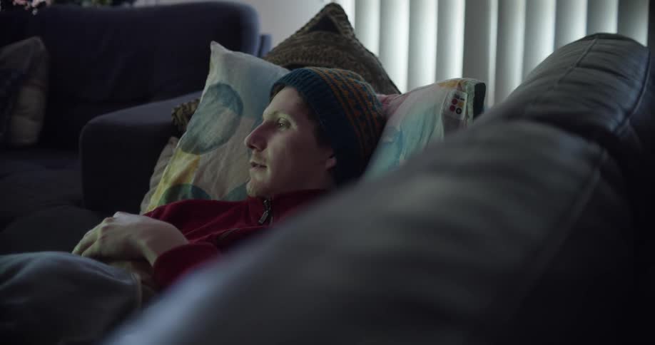 A man with a hat laying on a couch in a room with a wall and window in the background