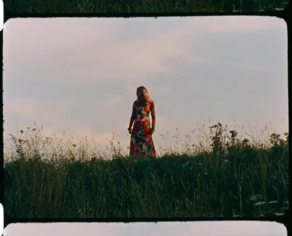 A woman with a dress standing in a landscape with grass with the sky and clouds in the background