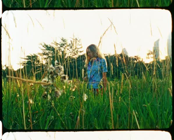 A woman standing in tall grass with wildflowers looking around and pulling back her blonde hair
