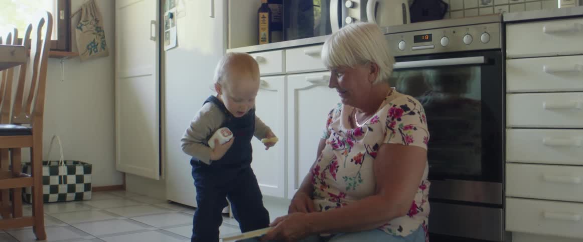 A woman sits on the floor in her kitchen and watches her grandson who walks over and looks at the book she is holding