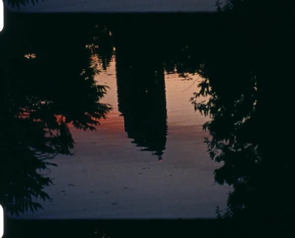 A tower and trees are reflected on the water surface at sunset