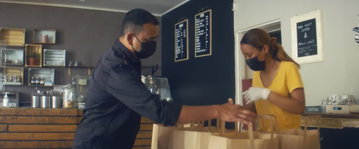 A woman with a mask and gloves putting styrofoam containers in brown paper bags as a man takes one of the bags