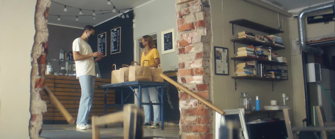 A woman packs foam food containers in a paper bag and gestures for a man to take one
