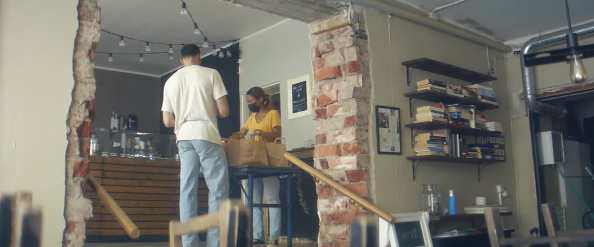 A woman arranging paper bags of food containers on a table is approached by a man who she allows to take a bag away