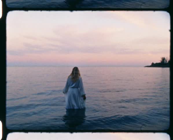 A woman in a gown stands in shallow water on the coast while gazing out at the horizon at sunset