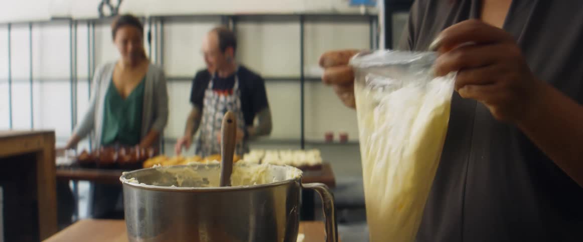 A woman fills an icing bag while two people behind her adjust cupcakes