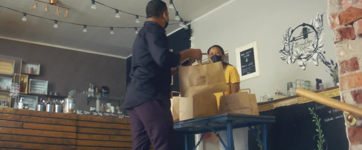 A woman with a mask and gloves standing by a table with brown paper bags as a man takes one of the bags