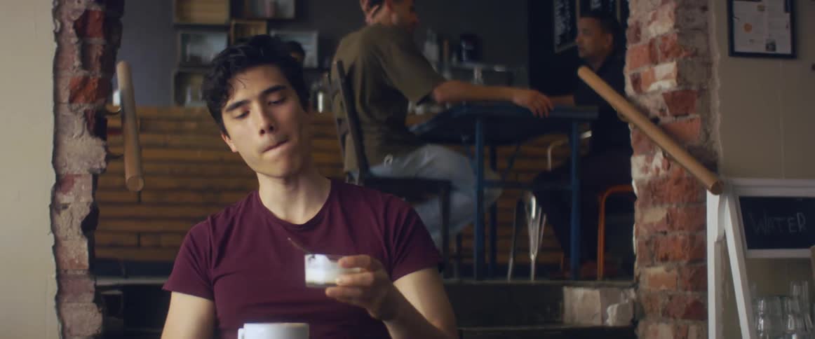 A young man eats a bowl of yogurt while sitting at a table in a cafe