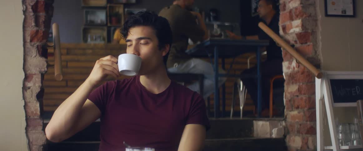 A young man sitting in a cafe takes a sip from his cup while chewing his food