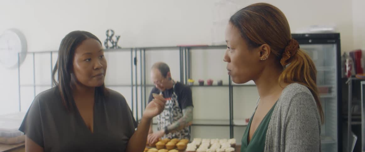 Two women talk and eat while a man adjusts cupcakes behind them