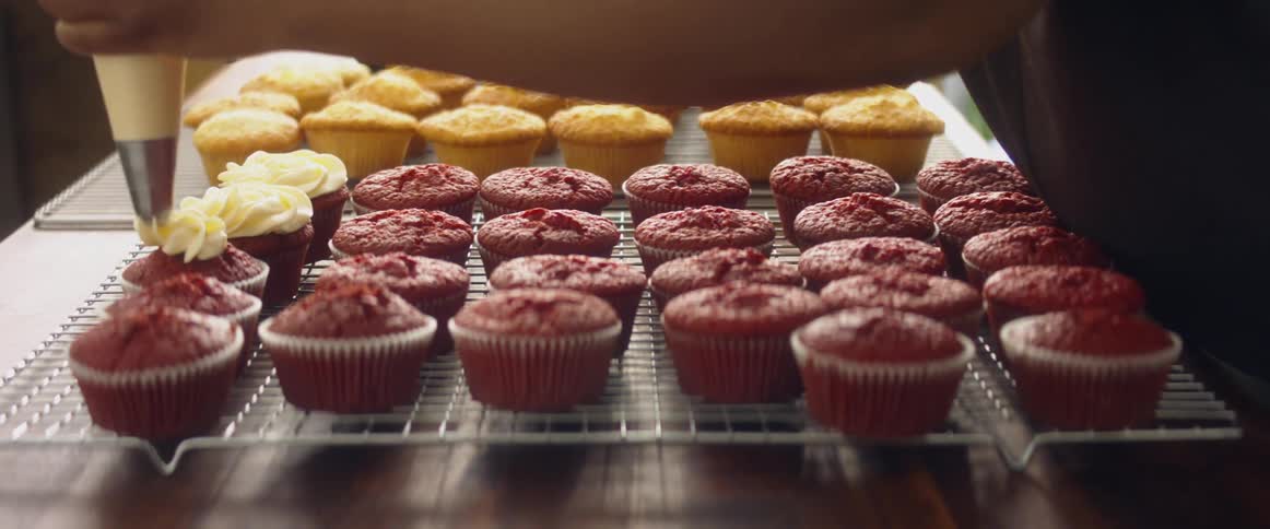 A baker frosts red velvet cupcakes on a wire rack