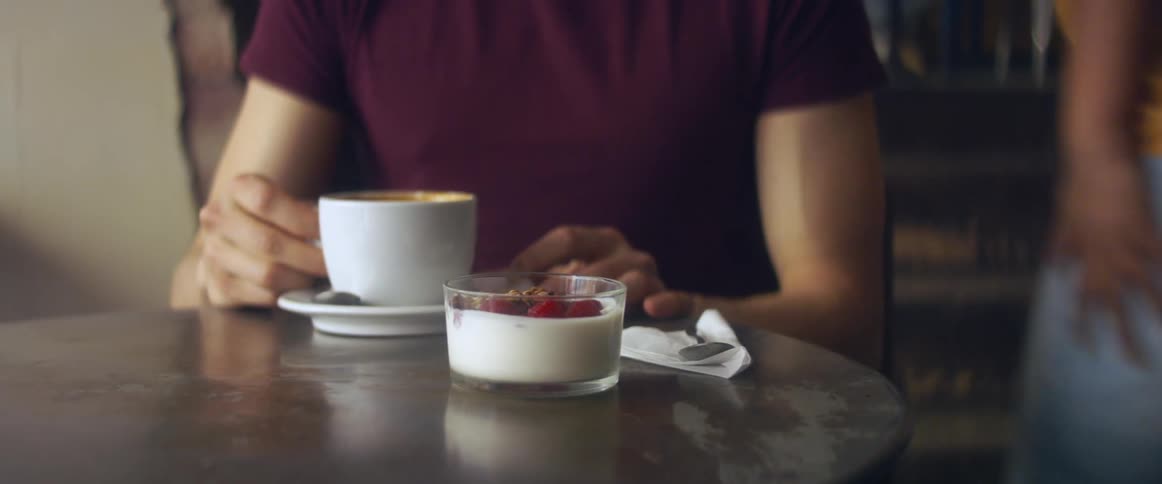 A young man sitting in a cafe drinks from a coffee mug after having his order brought to his table by a young woman