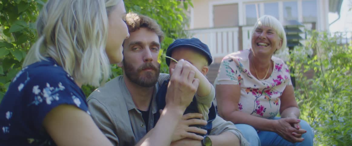 A boy sits on his fathers lap and holds a flower out for his mother and grandmother to smell