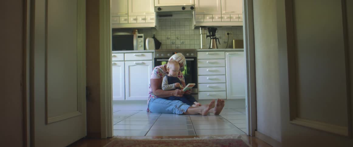 A woman sits down on the floor grabs a toddler boy on to her lap and starts to read a book to him
