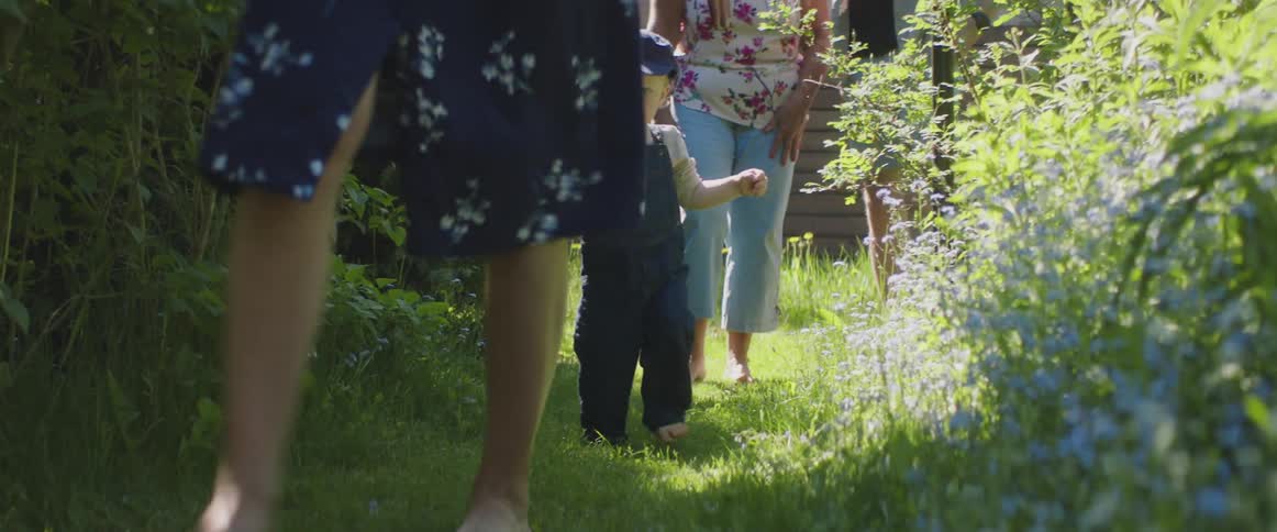 Two women a boy and a man walking on a path surrounded by plants with a house in the background